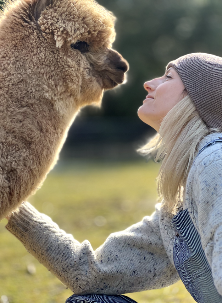 Amy smiling up at an affectionate alpaca, glowing in the sun